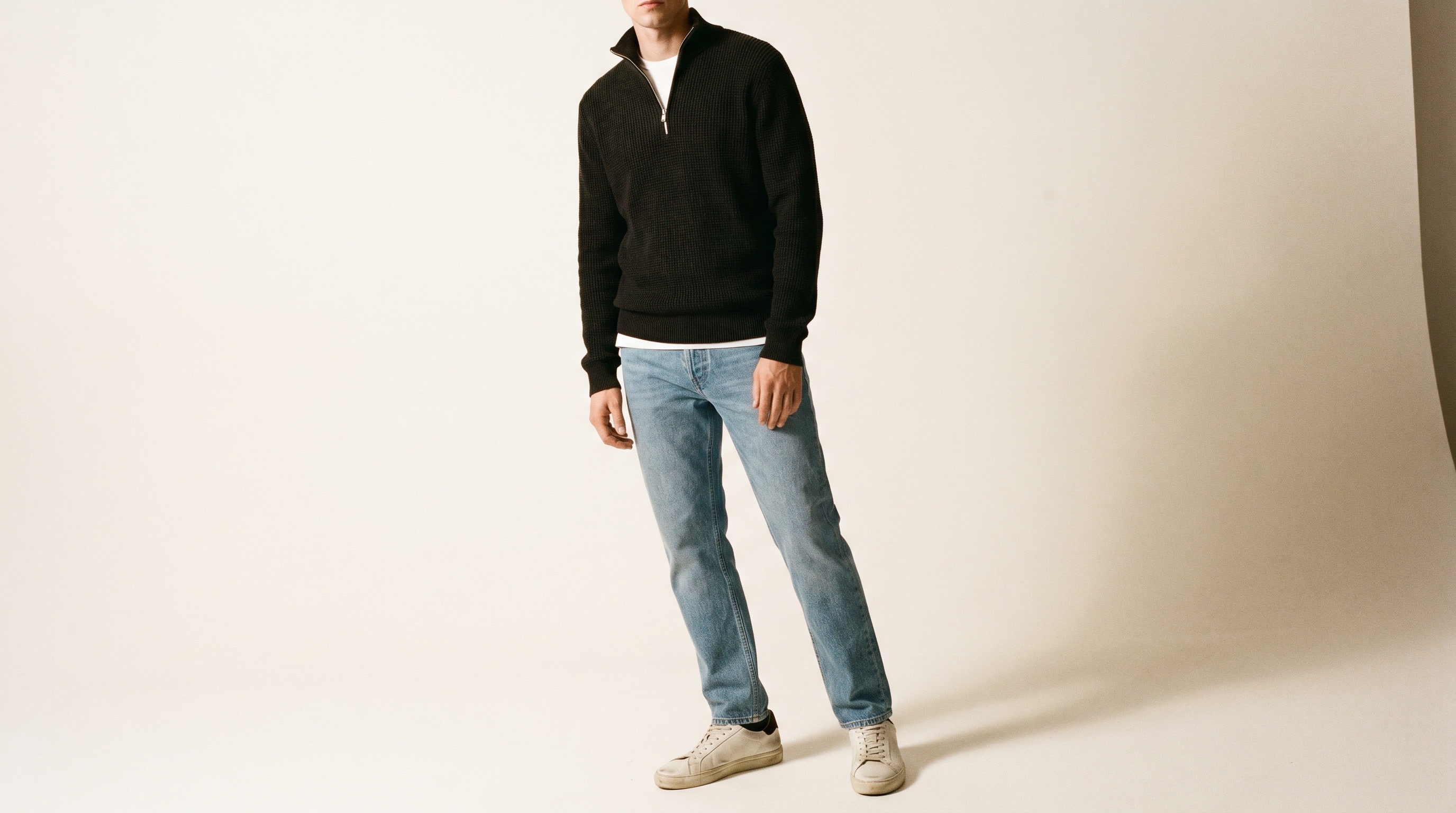Studio photo of a model wearing a black quarter-zip sweater, light wash jeans and white sneakers